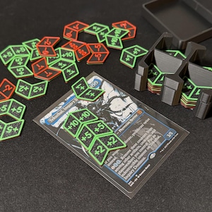 May include: A collection of gaming accessories, including dice-shaped counters with green and red accents, and black card holders. The counters display numbers like +5, +10, and -2. A card is also visible.