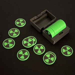 May include: A set of green and black game tokens with a radiation symbol, along with a matching gray storage box. The tokens are round and have a black center with a green radiation symbol. Some tokens are stacked inside the box.