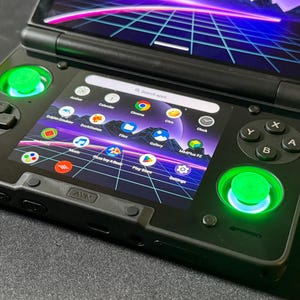 May include: Black handheld gaming device with a vibrant screen displaying app icons. The device features green illuminated joysticks and buttons. The screen shows a search bar and various app icons, including "Calendar" and "Play Store".