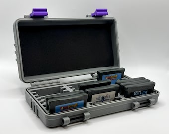 May include: Open grey plastic case with purple clasps, containing several video game cartridges. The case features a black foam interior and compartments for organising the game cartridges. The case is designed to protect and store video game cartridges.