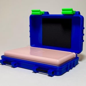 Puede incluir: Una funda protectora azul con cierres verdes, abierta para revelar una consola de juegos portátil rosa. La funda tiene un inserto de espuma negra y está diseñada para proteger el dispositivo. La funda mide aproximadamente 15 cm de largo.
