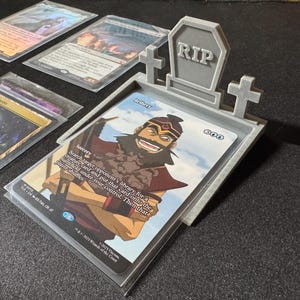 May include: A grey 3D-printed card holder in the shape of a tombstone, with the word "RIP" on the headstone. A card with the word "Bribery" is visible. Other cards are in the background. The card holder is designed to hold trading cards.