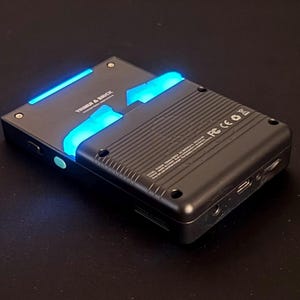 May include: Black electronic device with blue LED lighting. The device has the text "TRIMBLE & BRICK" on the top. The device has a ridged back and several ports. The device is on a dark surface.
