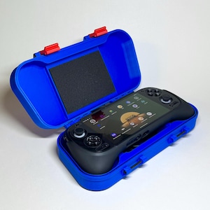 May include: A black handheld gaming console rests inside an open blue case with red latches. The console's screen displays an app interface. The case has a black foam insert. The case is on a white surface.