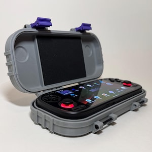 Puede incluir: Una funda gris impermeable con cierres morados, abierta para revelar un dispositivo de juego portátil negro. El dispositivo tiene una pantalla negra que muestra iconos y joysticks con acentos rojos. La funda está diseñada para proteger el dispositivo.