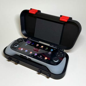 May include: A black handheld gaming console with a gray grip, displayed inside an open black case with red latches. The screen displays the text "Retro R" and various app icons. The case is on a white surface.