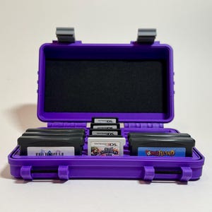 Puede incluir: Un estuche morado, abierto, de carcasa dura con un interior de espuma negra, que contiene varios cartuchos de videojuegos. El estuche está diseñado para guardar juegos de Nintendo DS y Game Boy Advance. El estuche tiene un asa y cierres.