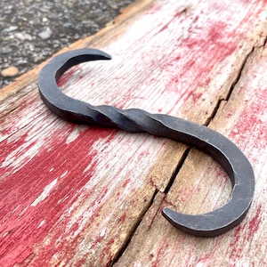 以下が含まれることがあります： 黒く仕上げられたねじれた金属製のフック。フックは金属製で、素朴な外観をしています。風化した木製の表面に置かれています。