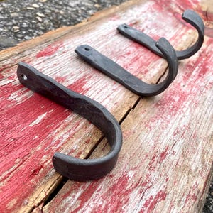 Puede incluir: Tres ganchos rústicos de metal negro montados en una tabla de madera desgastada. Los ganchos están forjados a mano y tienen una textura única y desigual.
