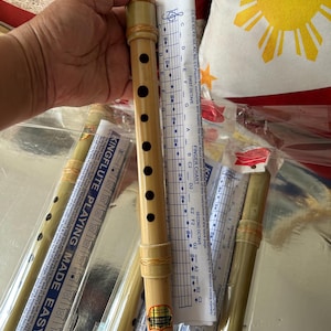May include: A light brown bamboo flute with seven black finger holes and decorative woven bands at the ends. The flute features a gold and red label near the bottom and is accompanied by a white instructional finger chart with blue text.