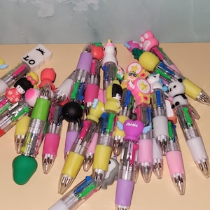 May include: A collection of colorful pens with various decorative toppers. The pens have multiple ink colors and are topped with designs such as a panda, a strawberry, a doll, and the word "Lonely". The pens are arranged on a light orange surface.