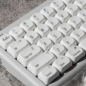 Puede incluir: Un teclado mecánico gris claro con teclas blancas redondeadas y letras negras. La tecla escape muestra la palabra hello en letra cursiva. El marco del teclado incluye el texto H05 y SYSTEM SETUP impreso en una fuente pequeña negra y roja.