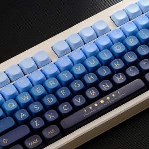 May include: A mechanical keyboard with a gradient blue keycap design. The keycaps transition from light blue to dark blue, with white lettering. The keyboard has a white frame and a dark blue spacebar with text and moon phase graphics.