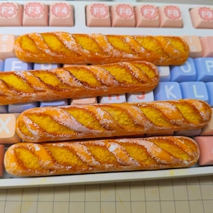 Könnte beinhalten: Vier Miniatur-Tastenkappen in Baguette-Form mit goldbrauner Kruste und gelber Innenstruktur, platziert auf einer mechanischen Tastatur mit pastellfarbenen rosa, blauen und weißen Tasten, die Buchstaben, Zahlen und F1- bis F10-Beschriftungen tragen.