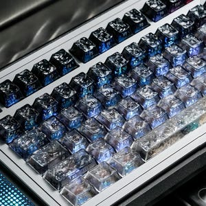 May include: A mechanical keyboard featuring custom keycaps with a translucent, dark blue and silver marbled resin finish. The keys display white lettering, including function keys and a spacebar with a textured, crystalline appearance.