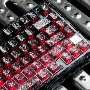 Puede incluir: Un teclado mecánico con teclas rojas translúcidas que presentan un patrón blanco fragmentado similar al cristal incrustado en cada una. El teclado tiene un marco de plástico transparente y una distribución estándar con teclas visibles.