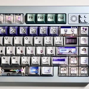May include: A mechanical keyboard with a light blue frame and custom keycaps. The keys feature white, purple, and dark green colours with various pixel art graphics, cat illustrations, and retro computer interface icons including Windows 98 text.