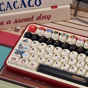 May include: A red and white keyboard with custom keycaps. The keycaps feature various designs, including Pokeball graphics and Japanese text. The keyboard also has the text "POKEMON GO!" on the spacebar.