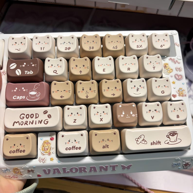 Puede incluir: Un teclado personalizado con teclas en forma de gato en tonos beige y marr&oacute;n. Las teclas presentan caras de gato e iconos. Algunas teclas tienen texto como "Tab", "Caps", "Good Morning", "coffee" y "alt". El teclado tiene una base azul claro con la palabra "VALORANTY".