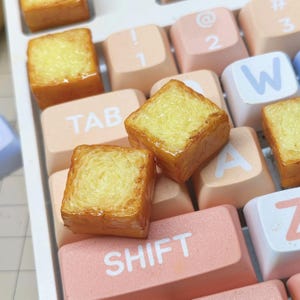 May include: Several miniature, square-shaped food items resembling toast are arranged on a keyboard. The toast is golden brown with a yellow, textured interior. The keyboard keys are various pastel colors, including pink, peach, and white.