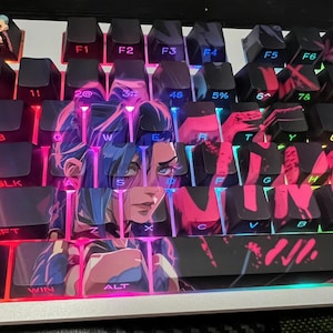 May include: A mechanical keyboard with custom keycaps featuring a character design in pink, blue, and purple. The keyboard has black keys with white lettering and a small figurine on the top left corner. The keyboard is lit with pink and blue lights.