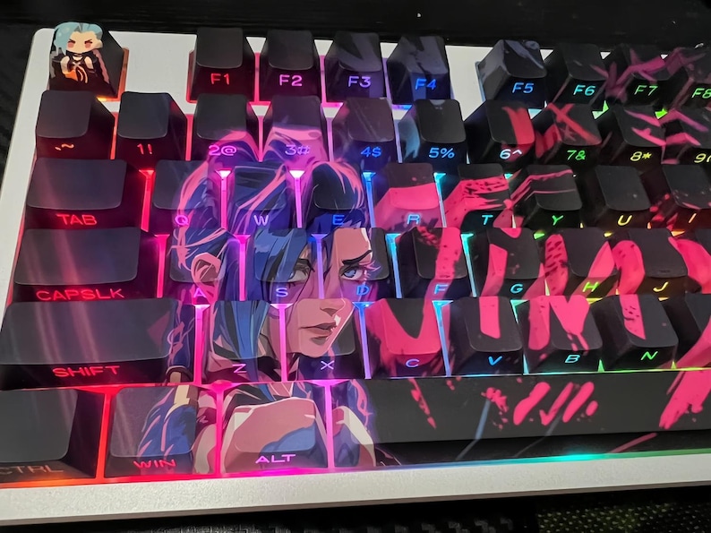 May include: A mechanical keyboard with custom keycaps featuring a character design in pink, blue, and purple. The keyboard has black keys with white lettering and a small figurine on the top left corner. The keyboard is lit with pink and blue lights.