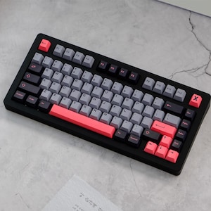May include: A black mechanical keyboard with gray and coral keycaps. The keyboard has a standard layout with function keys, a number pad, and arrow keys. The keycaps are a mix of gray and coral, with black keys for the function keys and modifiers.