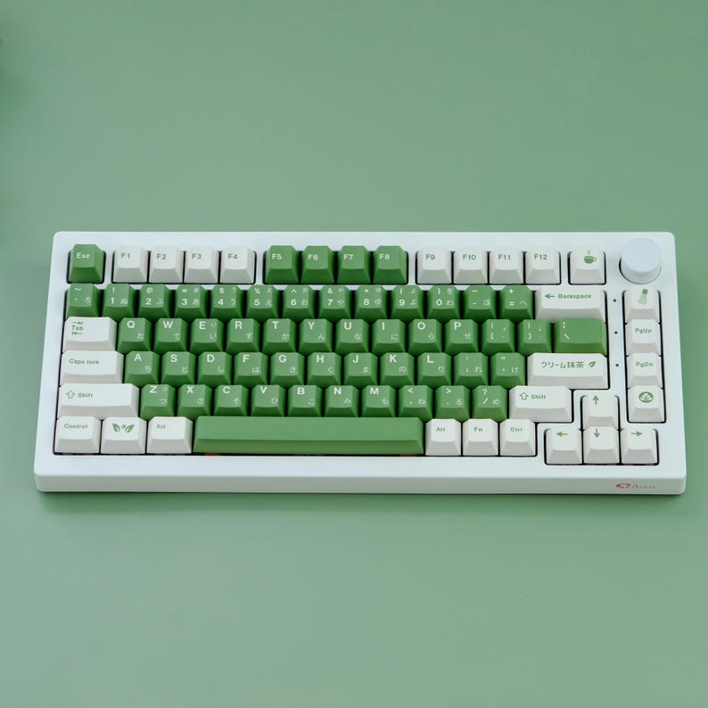 May include: A white mechanical keyboard with green and white keycaps. The keyboard has a full layout with function keys, a number pad, and a volume knob. The keycaps feature both English and Japanese characters. The keyboard sits on a green surface.