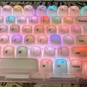 May include: A mechanical keyboard with translucent white rounded keycaps featuring black lettering. The keyboard is illuminated with vibrant multi-colored RGB backlighting, casting soft glows of pink, blue, orange, and purple beneath the keys.