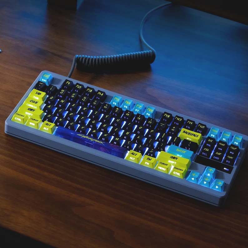 May include: A gray mechanical keyboard with yellow and blue keycaps. The keyboard has a wired connection and is set on a wooden surface. The keycaps have black lettering and the keyboard has a blue backlight.
