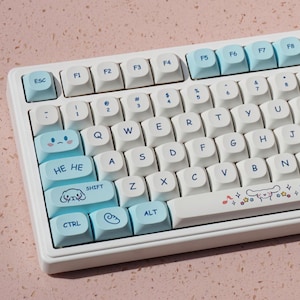 Può includere: Una tastiera meccanica bianca con tasti di accento azzurri. La tastiera presenta tasti personalizzati con grafica a cartoni animati e la scritta "HE HE". La tastiera ha un layout QWERTY standard.