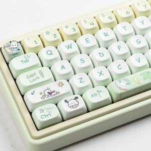 Può includere: Primo piano di una tastiera verde menta e bianca con tasti personalizzati. I tasti presentano illustrazioni di cartoni animati e testo. La tastiera ha una cornice verde chiaro e una base bianca. I tasti sono un mix di bianco e giallo chiaro.