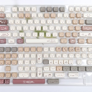 Puede incluir: Un conjunto de teclas de teclado en una bandeja de pl&aacute;stico transparente. Las teclas son principalmente blancas, beige y marrones, con dise&ntilde;os de caras de oso. Algunas teclas tienen gr&aacute;ficos tem&aacute;ticos de caf&eacute; y las palabras "Good Morning".