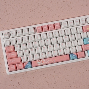 Puede incluir: Un teclado mecánico blanco con teclas rosas y azules. El teclado tiene teclas de función F1 a F12. Las teclas son blancas, rosas y azules, algunas con diseños decorativos. Fondo rosa.