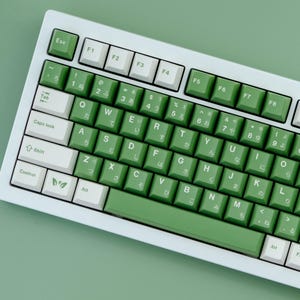 May include: A white and green mechanical keyboard with Japanese characters. The keyboard has white and green keycaps, with the function keys in white. The keys are arranged in a standard QWERTY layout.