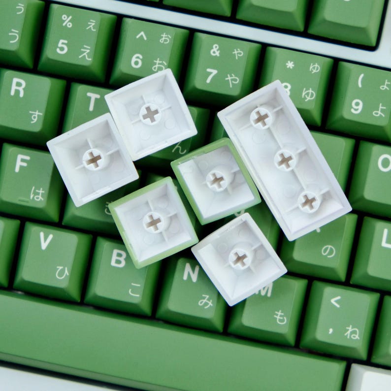 May include: Close-up of a green and white mechanical keyboard. The keyboard has green keycaps with Japanese characters and white keycaps scattered on top. The keycaps are square and have a cross-shaped design in the center.