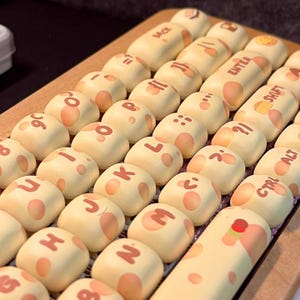 Puede incluir: Primer plano de un teclado con teclas personalizadas. Las teclas son de color crema con manchas marrones y tienen letras y símbolos impresos en ellas. El teclado está hecho de madera y tiene un acabado marrón.