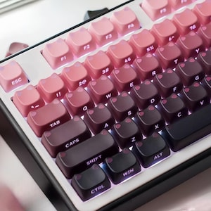 Op de afbeelding: Een mechanisch toetsenbord met katvormige keycaps in een gradiënt van roze en bruin. Het toetsenbord heeft een wit frame en een zwarte basis. De toetsen bevatten letters, cijfers en symbolen. De ESC-toets is roze en de CTRL-, WIN- en ALT-toetsen zijn zwart.