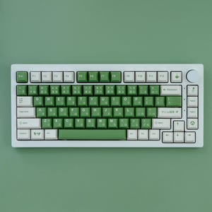 May include: A white mechanical keyboard with green and white keycaps. The keyboard has a compact layout with function keys and a rotary knob. The keycaps feature Japanese characters and English letters. The background is a solid green color.