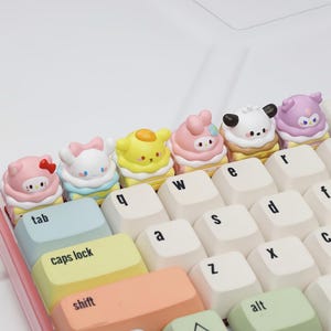 Pode incluir: Um teclado com teclas personalizadas com designs de personagens de desenhos animados coloridos. As teclas têm a forma de cupcakes com vários personagens em cima. O teclado tem uma moldura rosa e teclas brancas com letras e símbolos.