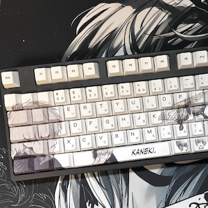 May include: A black and white keyboard with custom keycaps featuring anime-style illustrations. The keycaps have Japanese characters and English letters. The word "KANEKI" is printed on the spacebar.