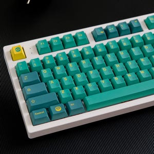 May include: A white mechanical keyboard with teal and yellow keycaps. The keyboard has a standard layout with function keys, number keys, and letter keys. The keycaps have a yellow outline and a teal background. The keyboard is on a black surface.