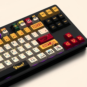 Puede incluir: Un teclado negro con teclas personalizadas que presentan personajes de Pok&eacute;mon. Las teclas son amarillas, rojas y blancas con varios dise&ntilde;os de Pok&eacute;mon. El teclado tiene una disposici&oacute;n japonesa con la palabra "Pokemon!" impresa en la barra espaciadora.