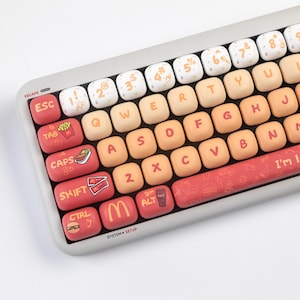May include: A white and gray mechanical keyboard with custom keycaps featuring fast food themed illustrations. The keycaps are in various shades of orange, red, and yellow. The keyboard has a row of keys labeled 'ESCAPE', 'ESC', 'TAB', 'CAPS', 'SHIFT', 'CTRL', 'ALT', and 'SYSTEM + SETUP'. The space bar has a red and white 'I'm lovin' it' graphic.