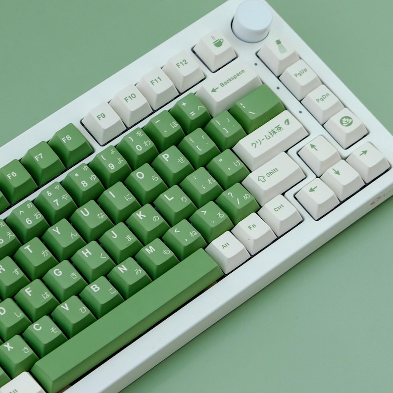May include: A white mechanical keyboard with green and white keycaps. The keycaps have Japanese characters and English letters. The keyboard has a volume knob and a variety of function keys. The color scheme is a light green and white.
