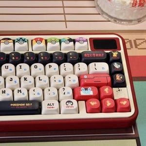 May include: A custom keyboard with a red and cream-colored frame. The keycaps feature a Pokemon theme, with various character designs and Pokeball icons. The keyboard includes Japanese text and the words "Pokemon Go!"
