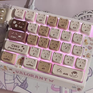 Puede incluir: Un teclado mec&aacute;nico blanco con teclas en forma de gato en tonos marr&oacute;n y crema. El teclado tiene el texto "GOOD MORNING" y "VALORANT". Las teclas presentan varios s&iacute;mbolos y letras.