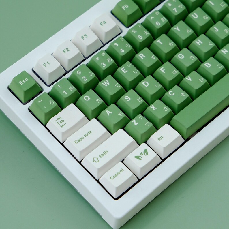 May include: A close-up of a white and green mechanical keyboard. The keyboard has white keys and green keys with Japanese characters. The keyboard is on a light green surface.