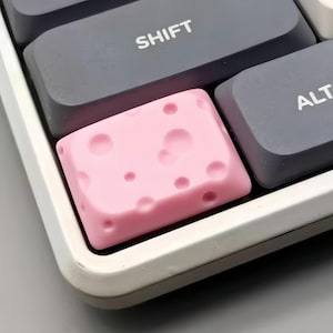 May include: A pink, cheese-shaped keycap with multiple holes, positioned between gray "SHIFT" and "ALT" keys on a white keyboard. The keycap adds a playful touch to a mechanical keyboard.
