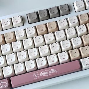 Puede incluir: Un teclado con teclas en forma de gato en tonos blanco, beige y gris. Las teclas presentan caras de gato y letras. La barra espaciadora es de color rosa empolvado y tiene el texto "COFFEE KITTY".